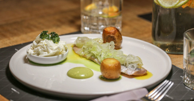 Na zdjęciu danie: kawałki ziemniaka, ryba i pojemnik z sosem, wszystko na białym talerzu. W tle szklanki i sztućce