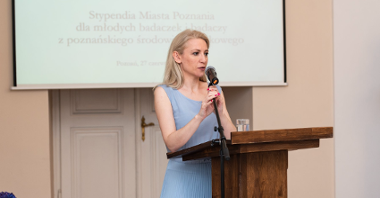 Na zdjęciu kobieta w błękitnej sukni, stoi przy mównicy z mikrofonem w ręku