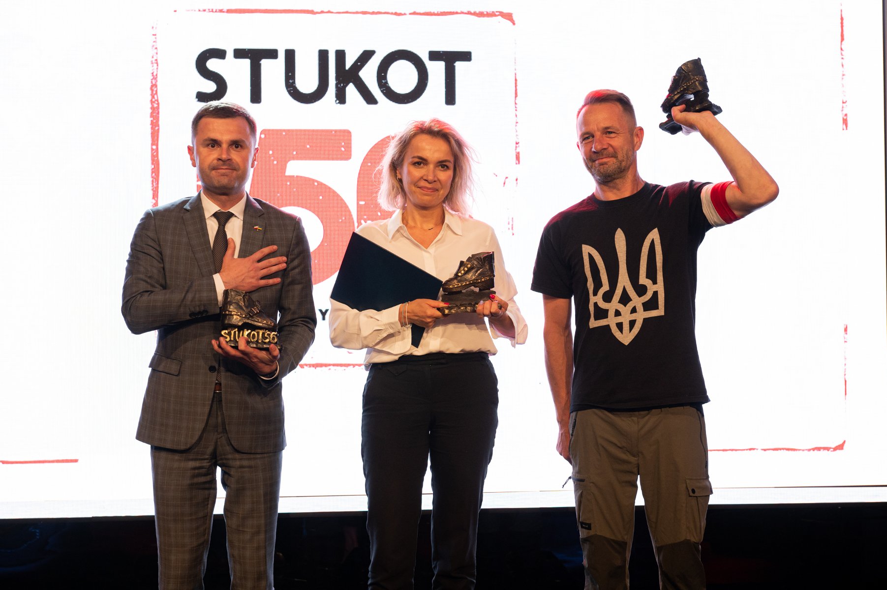 Na zdjęciu laureaci nagrody: dwaj mężczyźni i kobieta, wszyscy trzymają w rękach statuetki, w tle ekran z napisem Stukot '56 - grafika artykułu