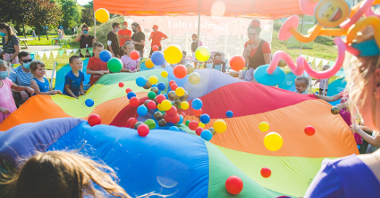 Zdjęcie przedstawia dzieci tłoczące się wokół wielkiej, kolorowej chusty klanza, na której leżą kolorowe piłki.