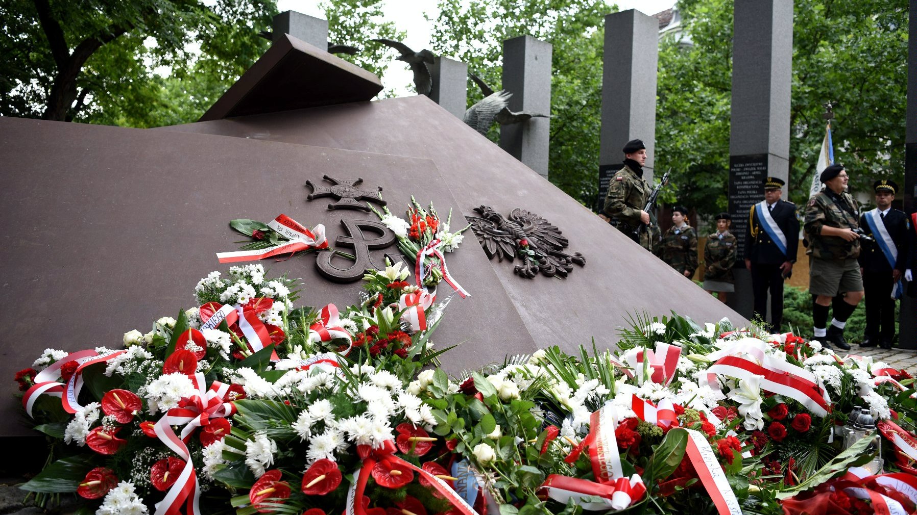 Na zdjęciu pomnik, pod nim wiązanki biało-czerwonych kwiatów - grafika artykułu