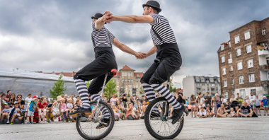 Na zdjęciu dwóch akrobatów na jednokołowych rowerach, trzymają się za ręce, widać też publiczność