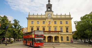 Na zdjęciu czerwony piętrowy autobus, stojący przed renesansowym ratuszem
