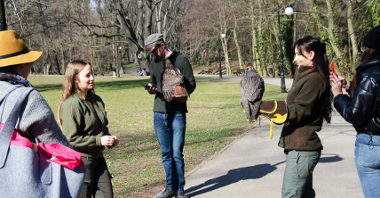Na zdjęciu ludzie w parku, dwójka z nich ma ptaki na rękach