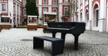 Galeria zdjęć przedstawiająca czarny fortepian ustawiony na dziedzińcu urzędu miasta, w tle budynki kolegiaty