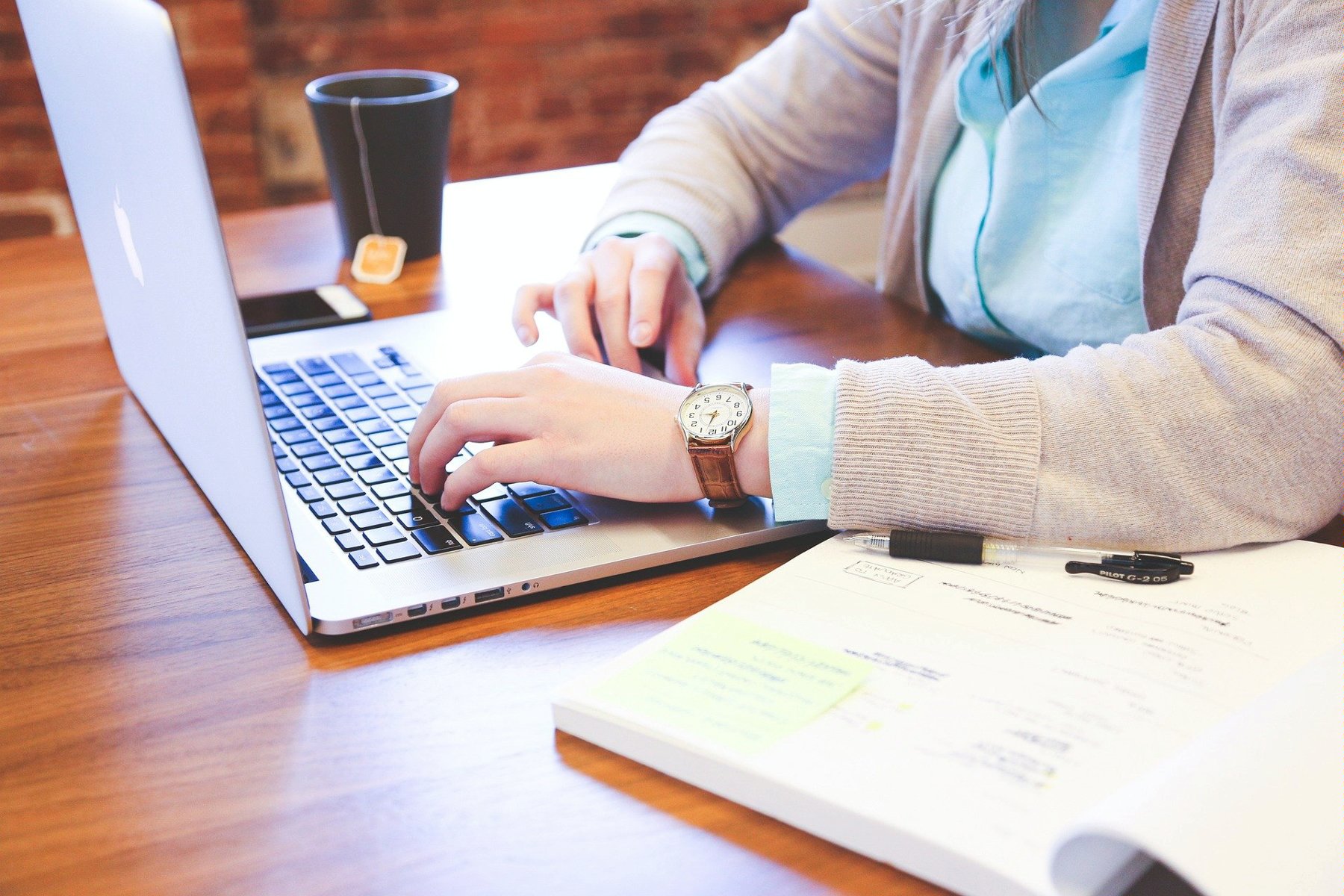Na zdjęciu osoba przed laptopem, na biurku stoi kubek oraz leżą papiery i telefon - grafika artykułu