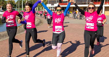 Na zdjęciu znajdują się cztery biegnące kobiety z podniesionymi rękoma, w tle więcej biegnących kobiet, wszystkie w różowych koszulkach