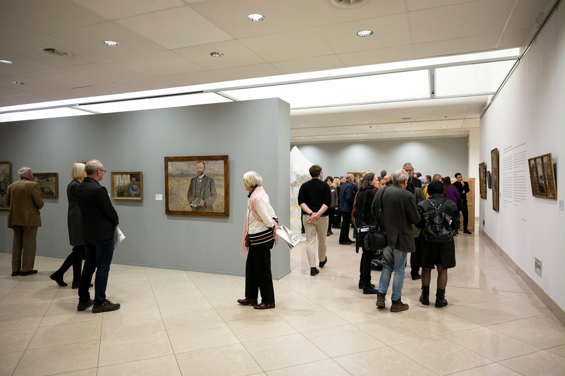 Grupa osób przechadza się po muzeum oglądając powieszone na ścianach obrazy. - grafika artykułu