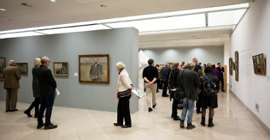 Grupa osób przechadza się po muzeum oglądając powieszone na ścianach obrazy.