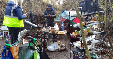 Na zdjęciu miejsce przebywania osób w kryzysie bezdomności. Widać półki z jedzeniem, namiot, stolik oraz osobę bezdomną