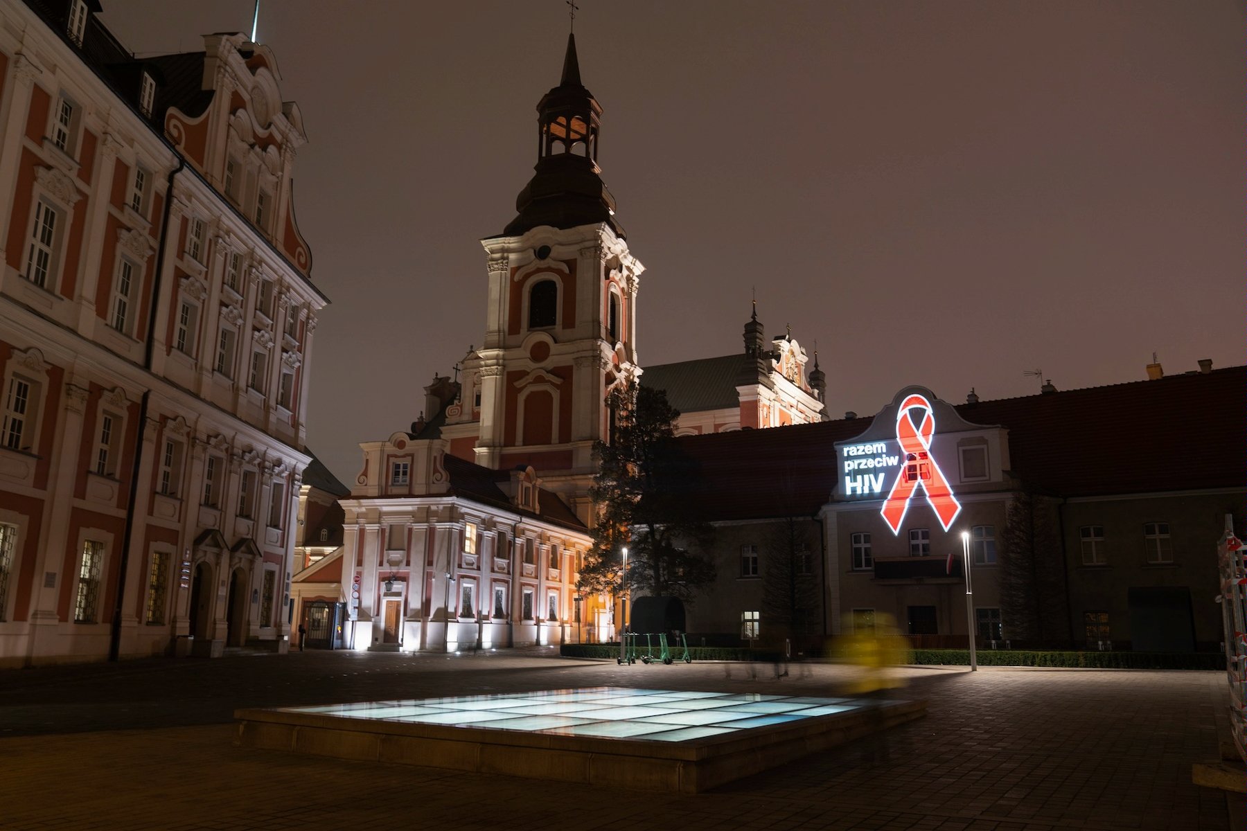 Na zdjęciu plac Kolegiacki, na jednym z budynków wyświetlona czerwona wstążeczka i napis: razem przeciw HIV - grafika artykułu