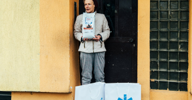 Na zdjęciu karmicielka, stoi w progu budynku, w dłoniach trzyma karmę