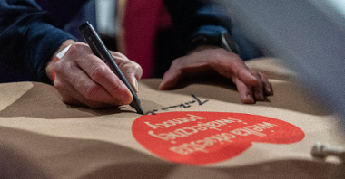 Na zdjęciu widać ręce, w jednej jest marker, osoba podpisuje bluzę z czerwonym serduszkiem WOŚP