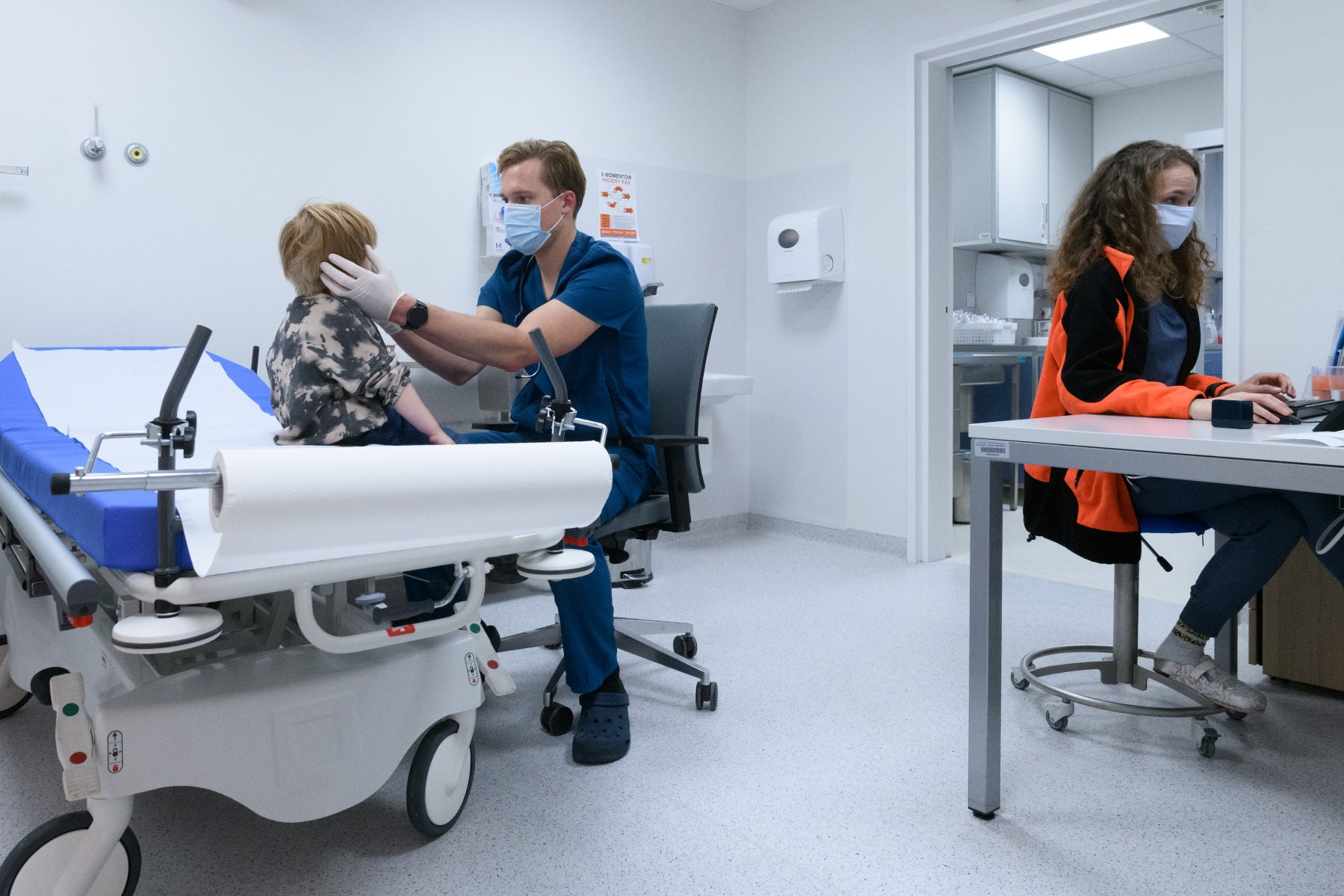 Zdjęcie przedstawia SOR w szpitalu na Wrzoska. Lekarz bada głowę dziecka. Przy biurku obok siedzi ratownik medyczny. - grafika artykułu