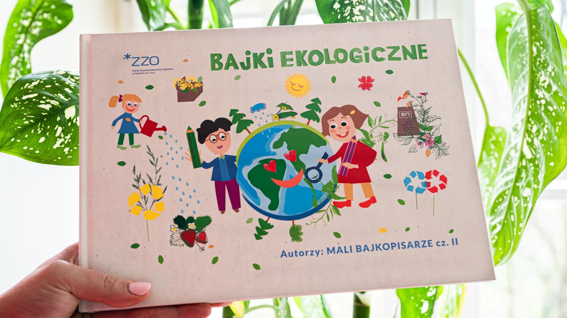 Na zdjęciu na tle zieleni okładka książki z dziećmi, kulą ziemską oraz elementami przyrody - grafika artykułu
