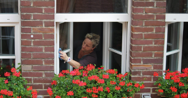 Na zdjęciu kobieta myjąca okno, przed nim kwitnące pelargonie