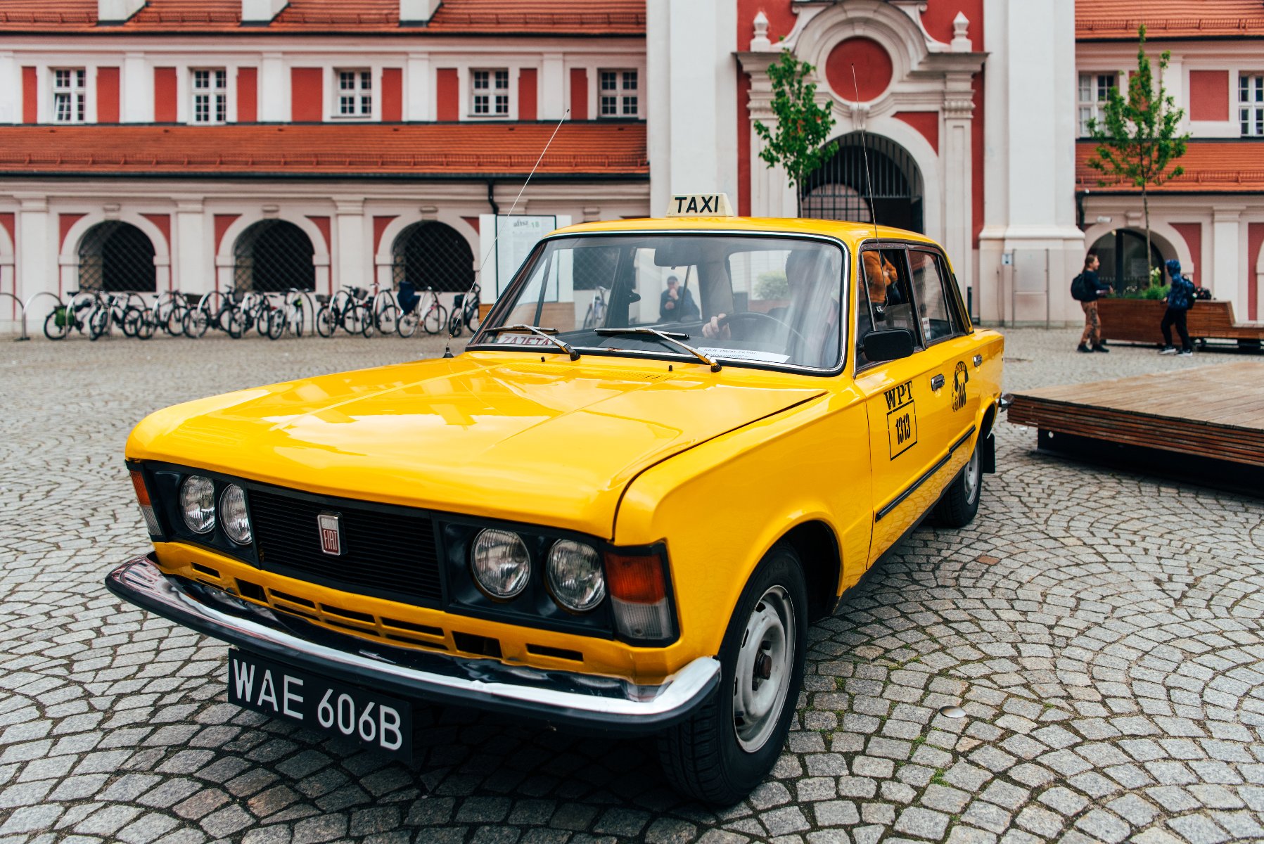 Na zdjęciu żółta starodawna taksówka na dziedzińcu urzędu miasta - grafika artykułu