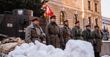 Zdjęcie przedstawia grupę rekonstruktorów w mundurach Powstania Wielkopolskiego. Za nimi stoi pojazd opancerzony.