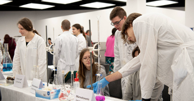 Zdjęcie przedstawia młodzież w białych fartuchach za stołem laboratoryjnym.