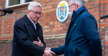 Na zdjęciu dwóch mężczyzn podających sobie ręce, jednym z nich jest prezydent Poznania, za nimi ceglana ściana, na niej herb Miasta