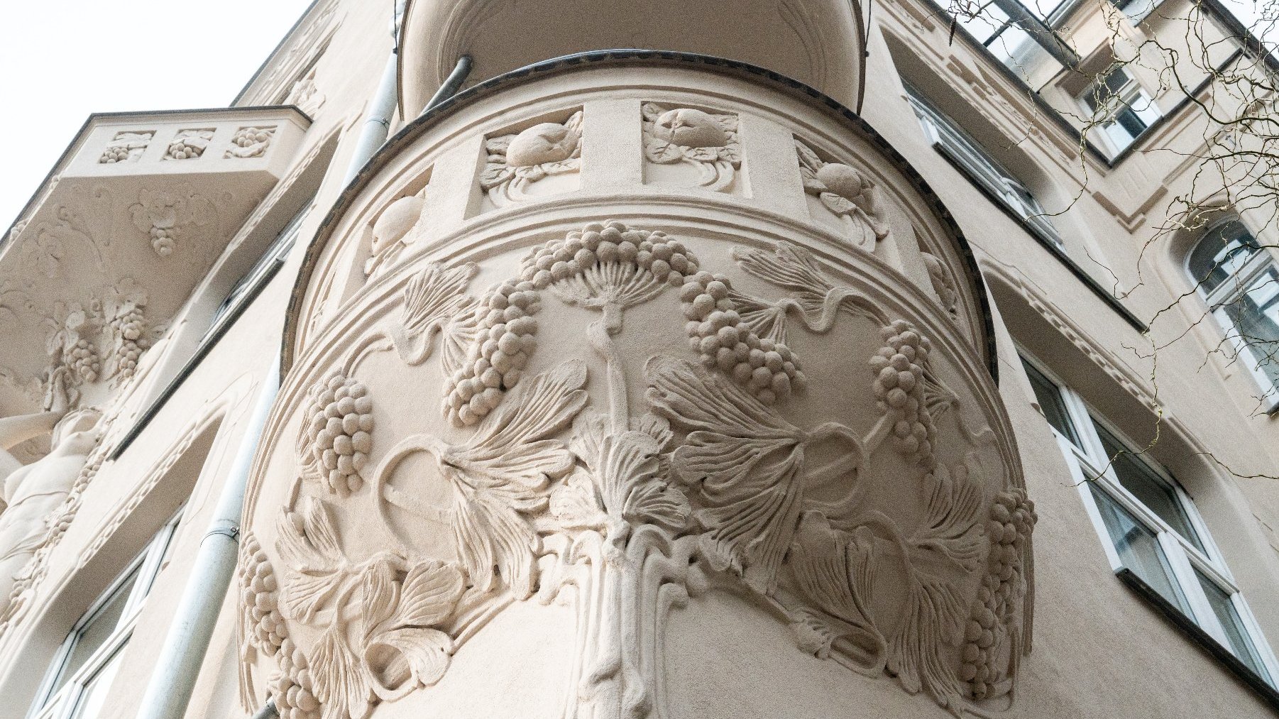 Zewnętrzna część wielopiętrowego budynku o misternych detalach architektonicznych. Budynek posiada ozdobne rzeźbienia i dekoracje, w tym wyróżniający się zaokrąglony balkon z detalami w formie winorośli i liści. Fasada zawiera również inne dekoracyjne elementy, takie jak rzeźbione postacie i wzory. Budynek wydaje się mieć kilka pięter, z licznymi oknami i jasną elewacją.