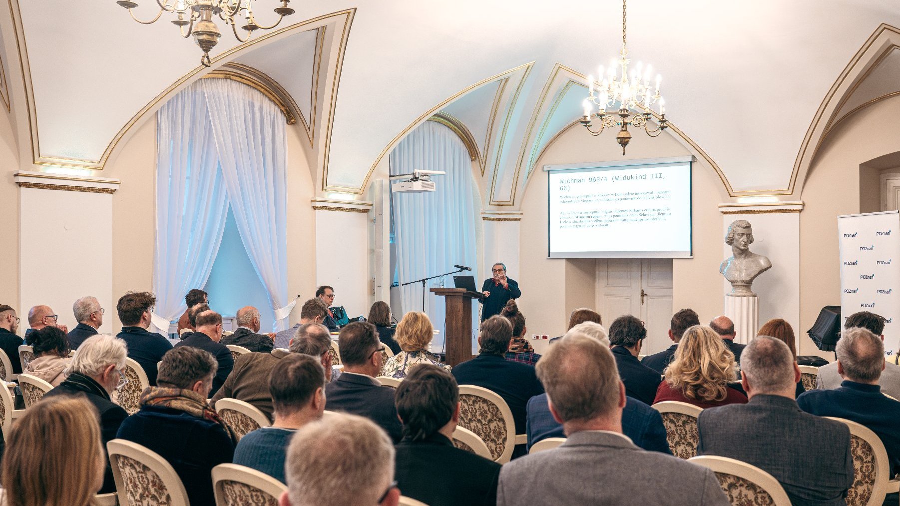 Zdjęcie przedstawia mężczyzne przy mównicy oraz tłum ludzi siedzących na sali.
