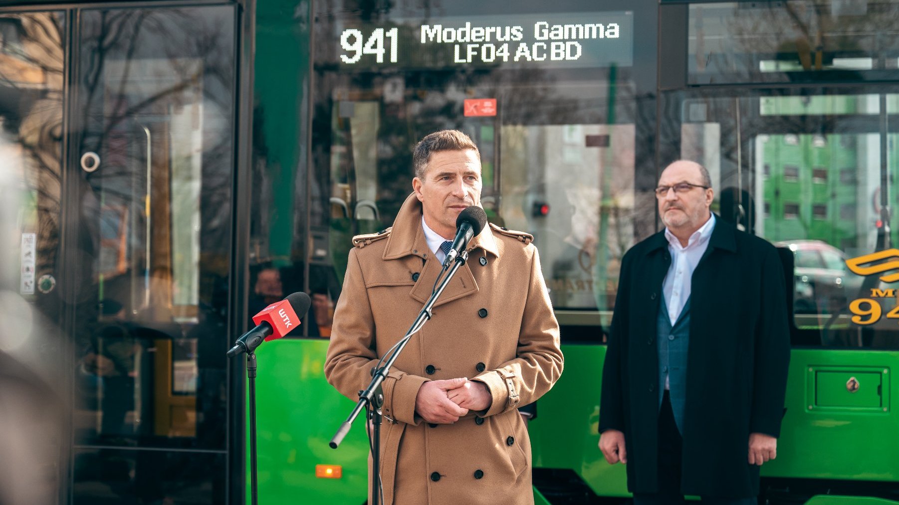 Na zdjęciu dwóch mężczyzn, jeden mówi coś do mikrofonu, za nimi tramwaj