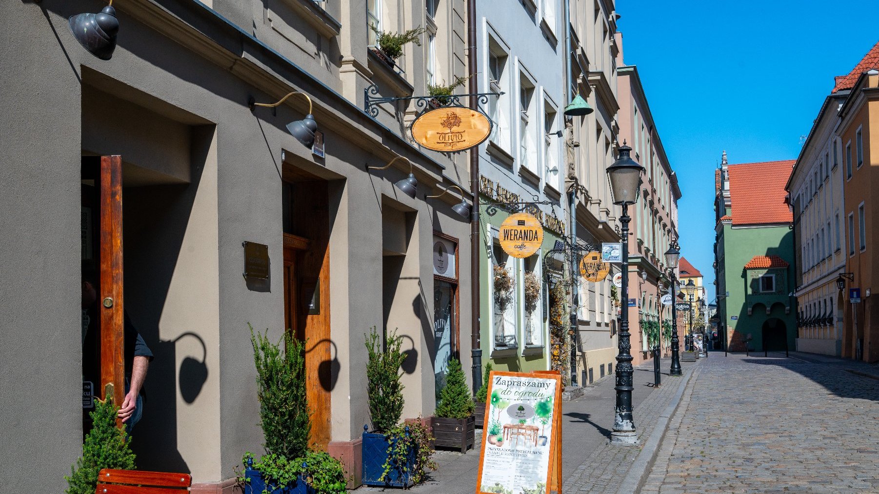 Ulica w europejskim mieście. Wzdłuż brukowanej ulicy stoją budynki z kawiarniami i sklepami na parterze. Na szyldach widoczne napisy: "Weranda cafe" i "Zapraszamy do ogrodu".
