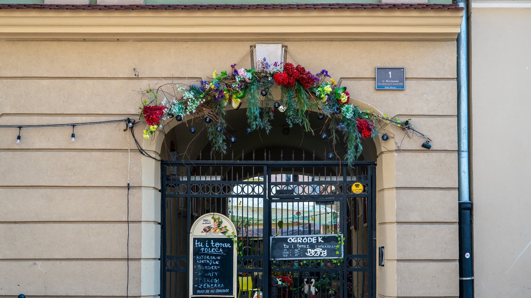 Wejście do budynku z ozdobnym łukiem pełnym kwiatów i zieleni. Czarne metalowe drzwi z misternymi zdobieniami prowadzą do wnętrza. Nad wejściem widnieje szyld z napisem w języku polskim.