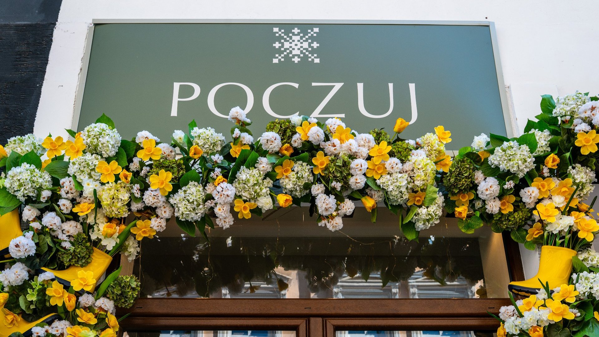 Sklep o nazwie "POCZUJ" z dekoracyjnym szyldem. Nad wejściem widnieje zielony napis "POCZUJ" na tle kwiatowej girlandy złożonej głównie z białych i żółtych kwiatów, w tym żonkili i hortensji. Dodatkowym elementem ozdobnym jest pikselowy wzór przypominający śnieżynkę. Girlanda otacza górną część witryny sklepowej, tworząc ciepłą i zapraszającą aranżację.