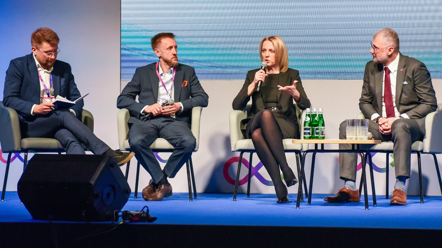 Na zdjęciu trzech mężczyzn i kobieta rozmawiają ze sobą, siedząc na krzesłach, kobieta trzyma mikrofon