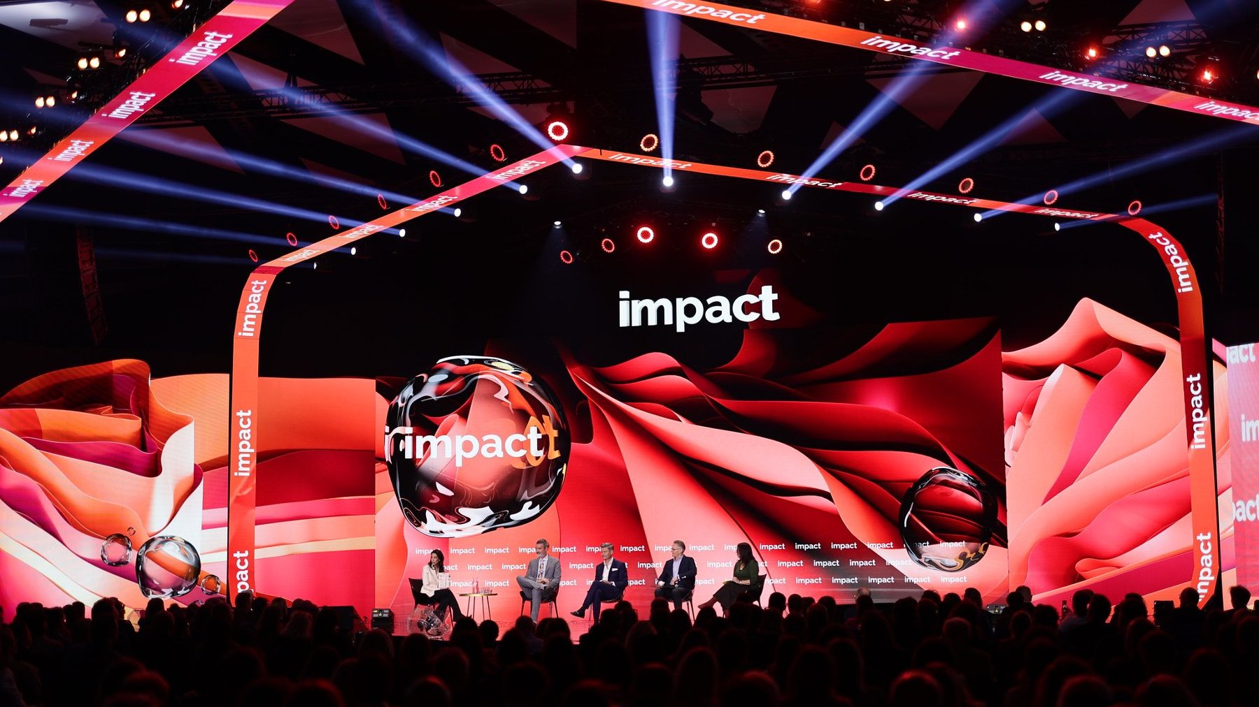 Scena konferencyjna z dużym ekranem wyświetlającym wielokrotnie słowo "impact". Na scenie pięć osób siedzących na krzesłach, prawdopodobnie uczestniczących w panelu dyskusyjnym. Tło w odcieniach czerwieni i pomarańczu, kilka kulistych obiektów także z napisem "impact". Dynamiczne oświetlenie skierowane na scenę, w tle widoczna publiczność