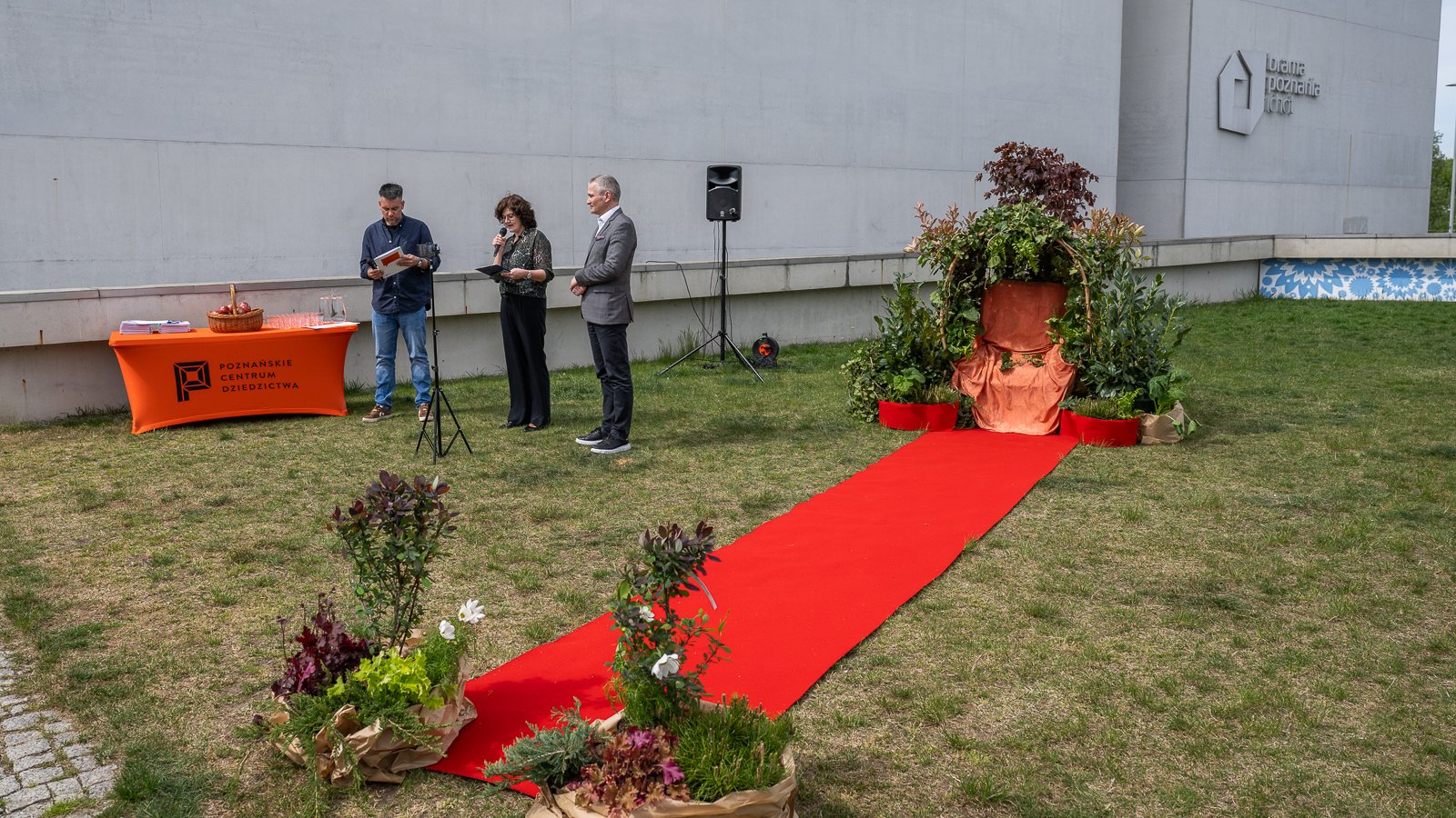 Zdjęcie przedstawiadwóch mężczyzn i kobietę na tle zieleni. Trzymają oni kartki papieru i mikrofony. Obok nich znajduje się czerwony dywan oraz instalacja z roślin, a także stół, na którym stoi koszyk z jabłkami.