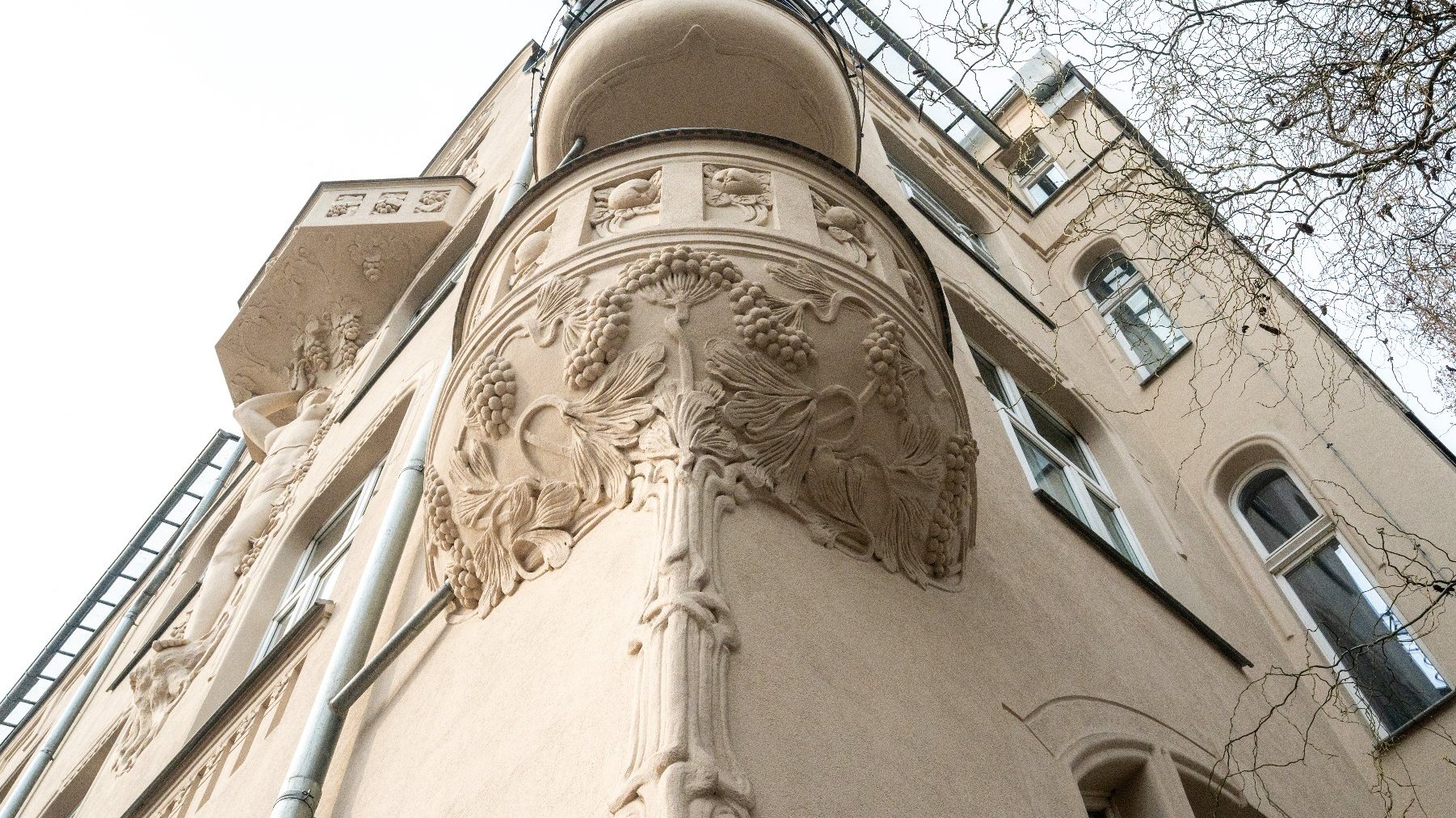 Zewnętrzna fasada budynku z bogato zdobionymi detalami architektonicznymi. Na zaokrąglonym narożniku znajduje się ornamentyka, w tym motywy roślinne przypominające winorośl. Budynek ma kilka okien, a fotografia została wykonana z perspektywy patrzenia w górę.