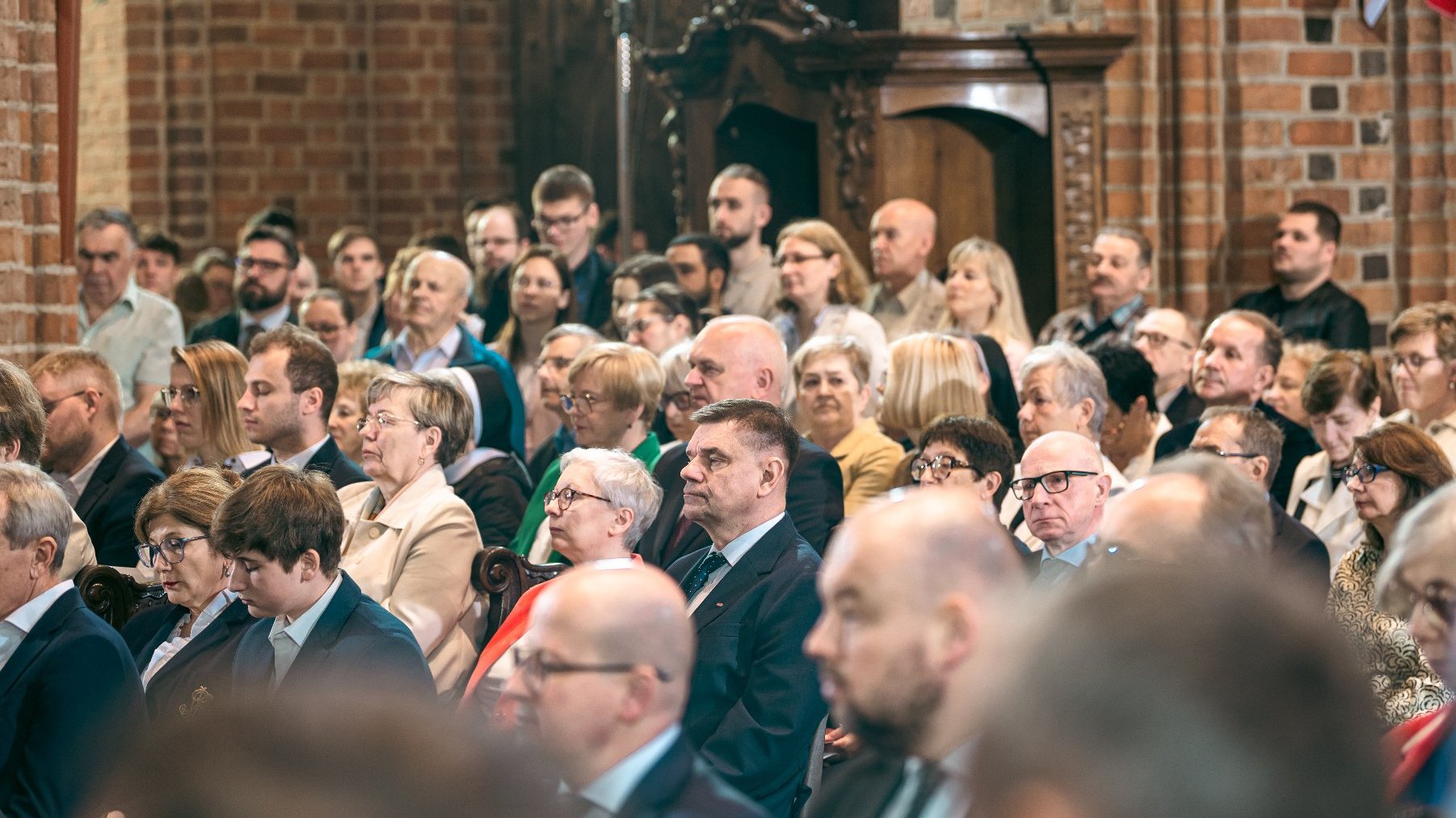 Uroczyste wydarzenie w pomieszczeniu z ceglaną ścianą w tle i elementami architektonicznymi z drewna. Na sali widoczna jest duża liczba osób ubranych formalnie, w garnitury i eleganckie sukienki, co sugeruje oficjalny charakter spotkania.