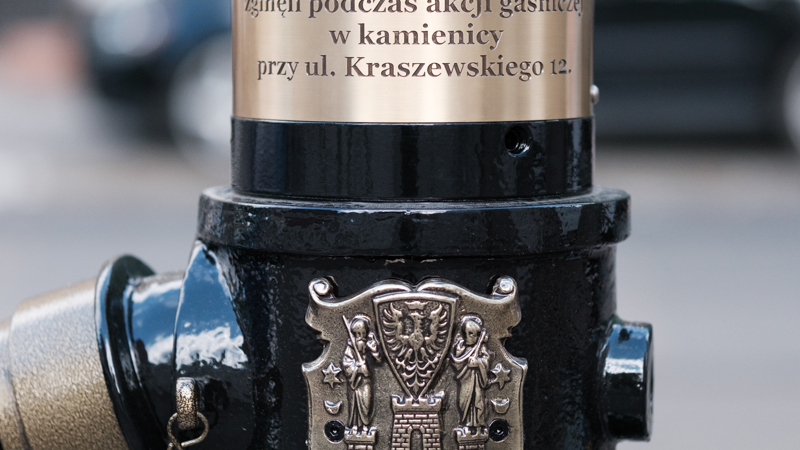Zdjęcie przedstawia tabliczkę upamiętniającą zmarłych strażaków umieszczoną na hydrancie wraz z herbem miasta.