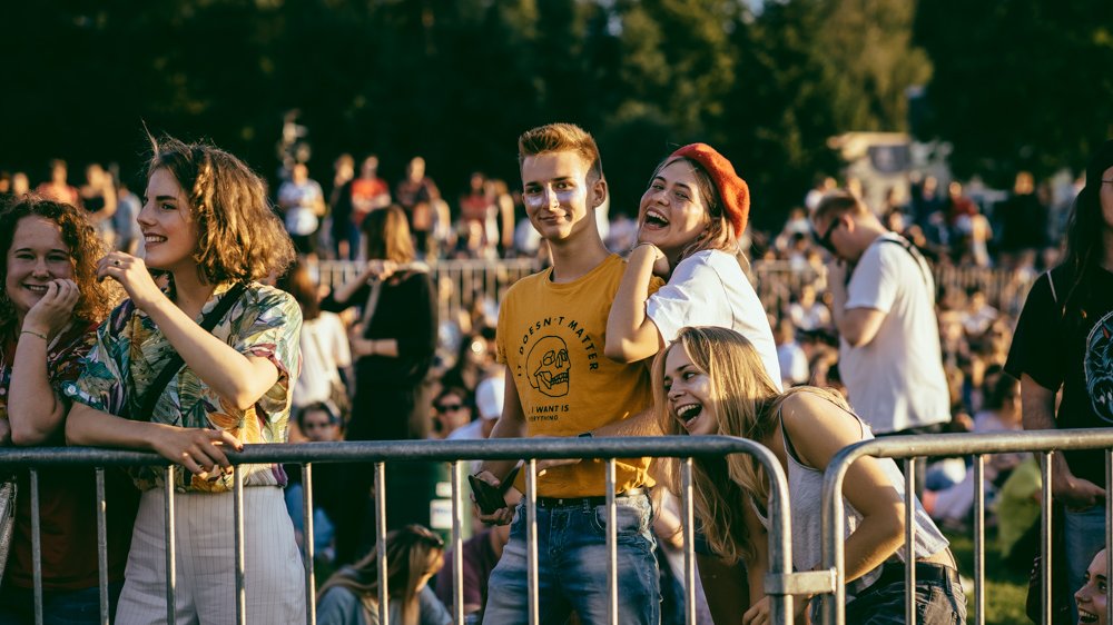 Grupa ludzi stojąca za metalową barierką na plenerowym wydarzeniu. Teren zielony, w tle drzewa. Uczestnicy ubrani są w codzienne stroje, w tym krótkie spodenki, koszulki i czapki.