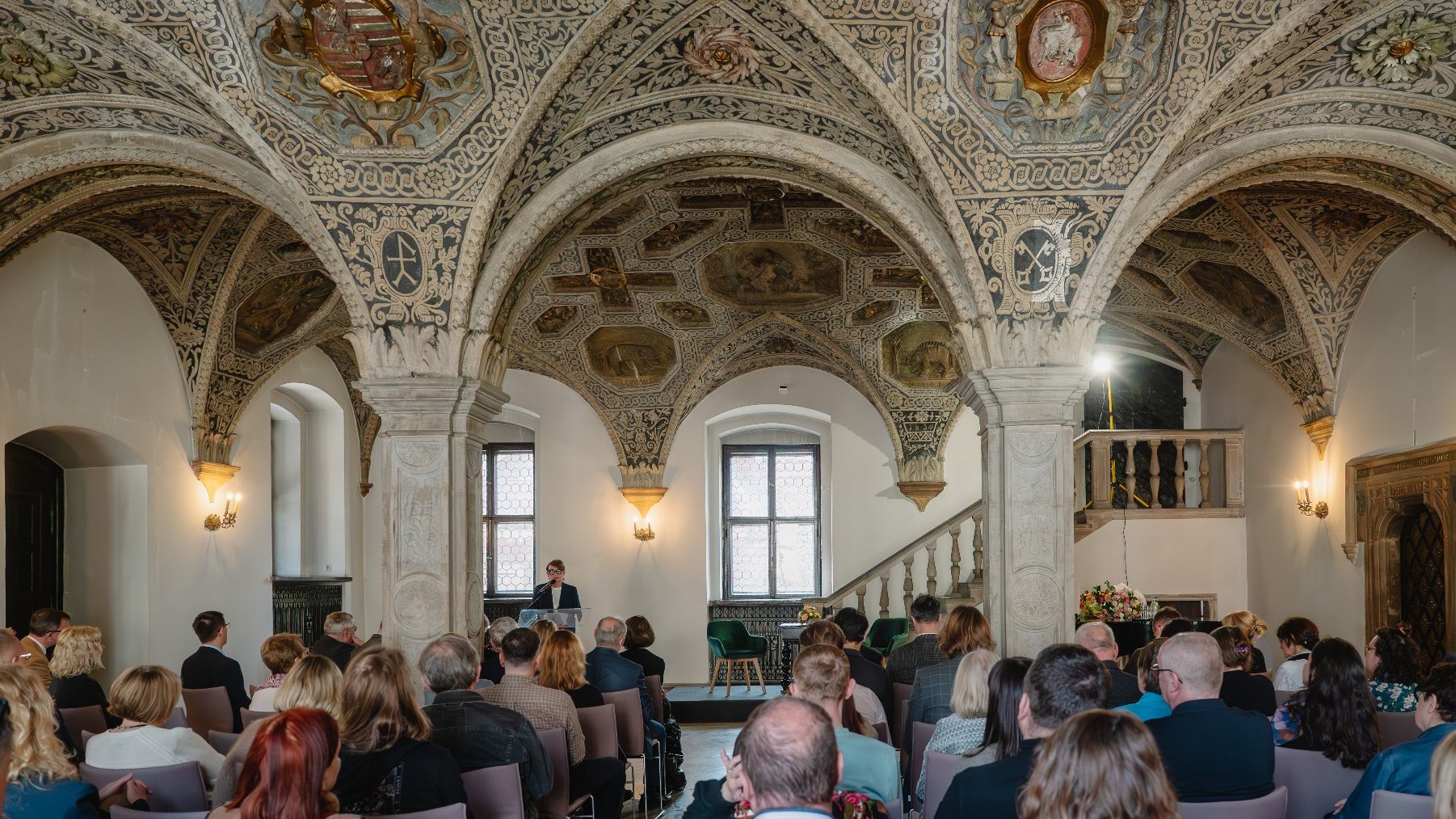 Zdjęcie przedstawia kobietę mówiącą do mikrofonu na mównicy i słuchający ją tłum ludzi w sali.