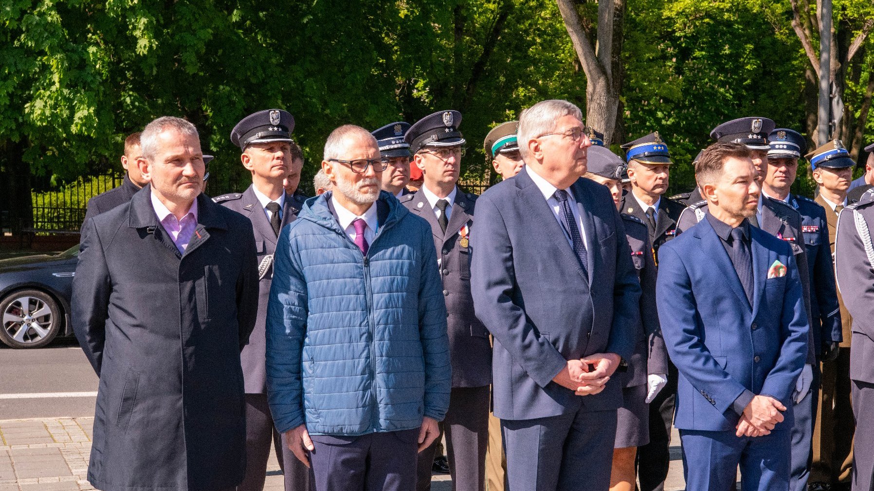 Grupa osób stoi na świeżym powietrzu podczas formalnego wydarzenia. Są ubrani w eleganckie stroje, w tym garnitury, krawaty i mundury. Niektóre mundury mają odznaczenia, medale i czapki, sugerujące obecność policji lub wojska.