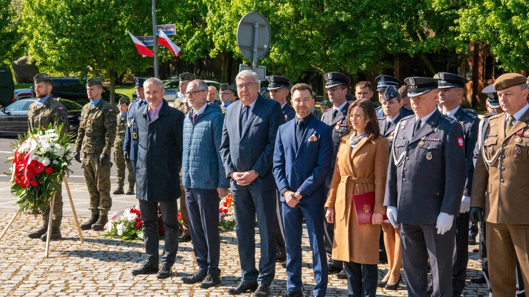 Grupa osób stoi na świeżym powietrzu podczas uroczystości. Wśród nich znajdują się osoby w mundurach wojskowych i policyjnych oraz w eleganckich strojach cywilnych. Na pierwszym planie leżą dwa wieńce z czerwonymi i białymi kwiatami.