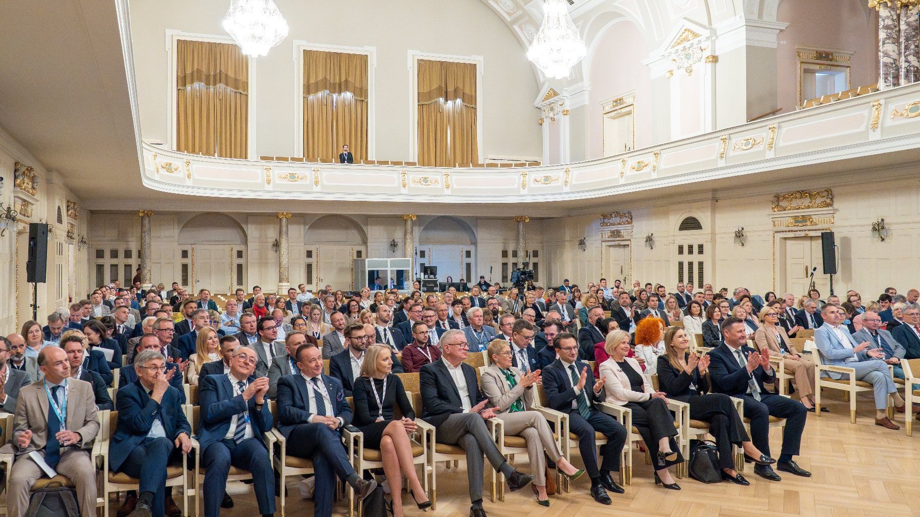 Publiczność siedzi w rzędach krzeseł, skierowana ku przodowi sali, a część osób bije brawo.