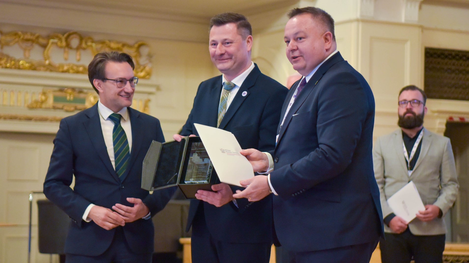 Cztery osoby w eleganckich strojach stoją w ozdobnym wnętrzu o bogato zdobionych ścianach. Trzy osoby trzymają dokument oraz szklaną statuetkę, co sugeruje oficjalne wręczenie nagrody lub wyróżnienia.
