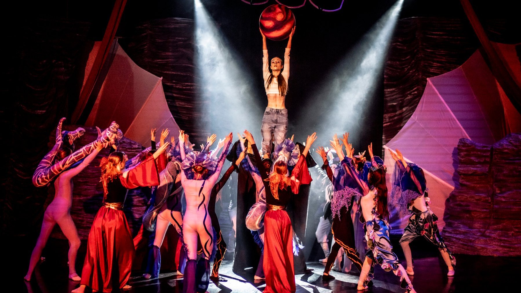 Scena teatralna z przedstawienia "Alia". W centrum kobieta unosi nad głową czerwoną, kulistą kulę, oświetlona dwoma reflektorami. Dookoła niej grupa aktorów w kolorowych, artystycznych strojach z uniesionymi rękami.