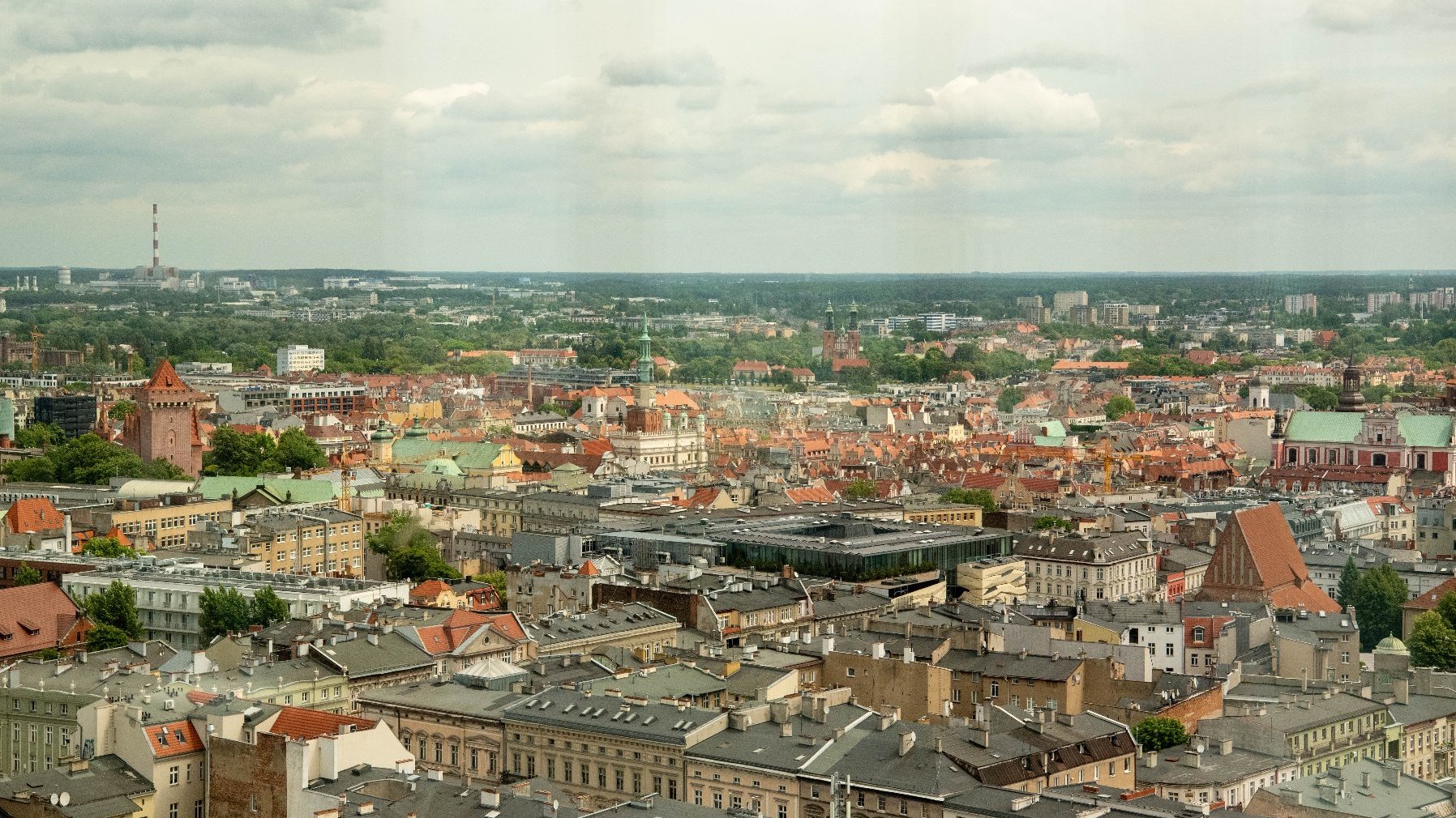 Panorama Poznania, widziana z wysokości, z licznymi dachami budynków. W oddali widoczne są wieże kościołów, zamek, a na horyzoncie komin elektrowni. Niebo jest pochmurne.