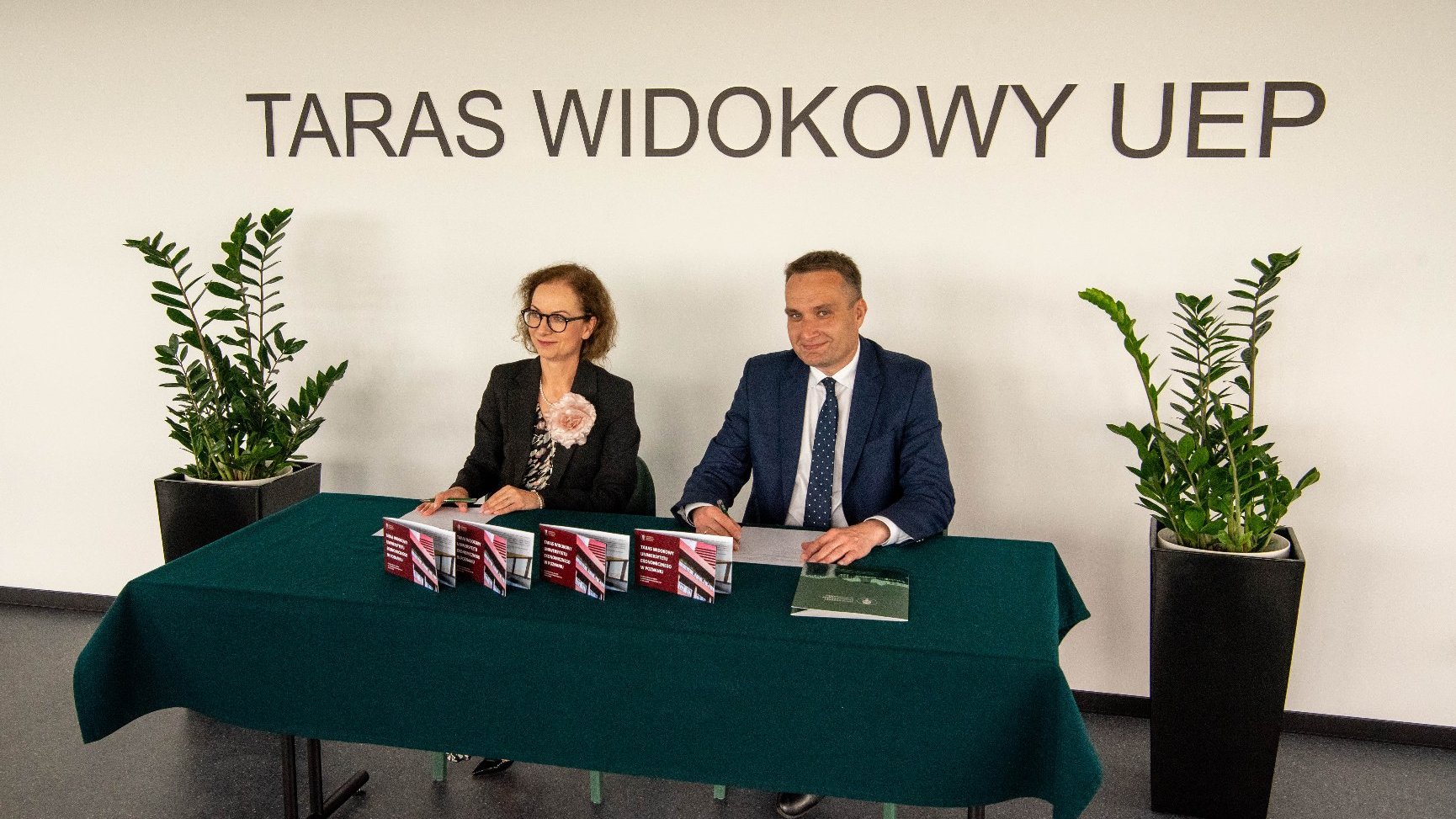 Kobieta w okularach i mężczyzna w garniturze siedzą za stołem nakrytym ciemnozielonym obrusem. Na stole leżą broszury z napisem "Taras Widokowy UEP". Za nimi biała ściana z dużym czarnym napisem "TARAS WIDOKOWY UEP" i dwoma zielonymi roślinami w donicach.