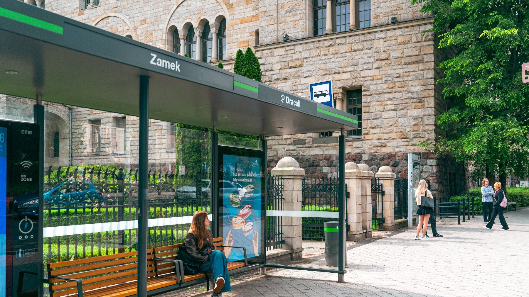 Zdjęcie przedstawia przystanek tramwajowy z napisem Zamek Draculi. Pod wiatą, na ławce siedzi dziewczyna.