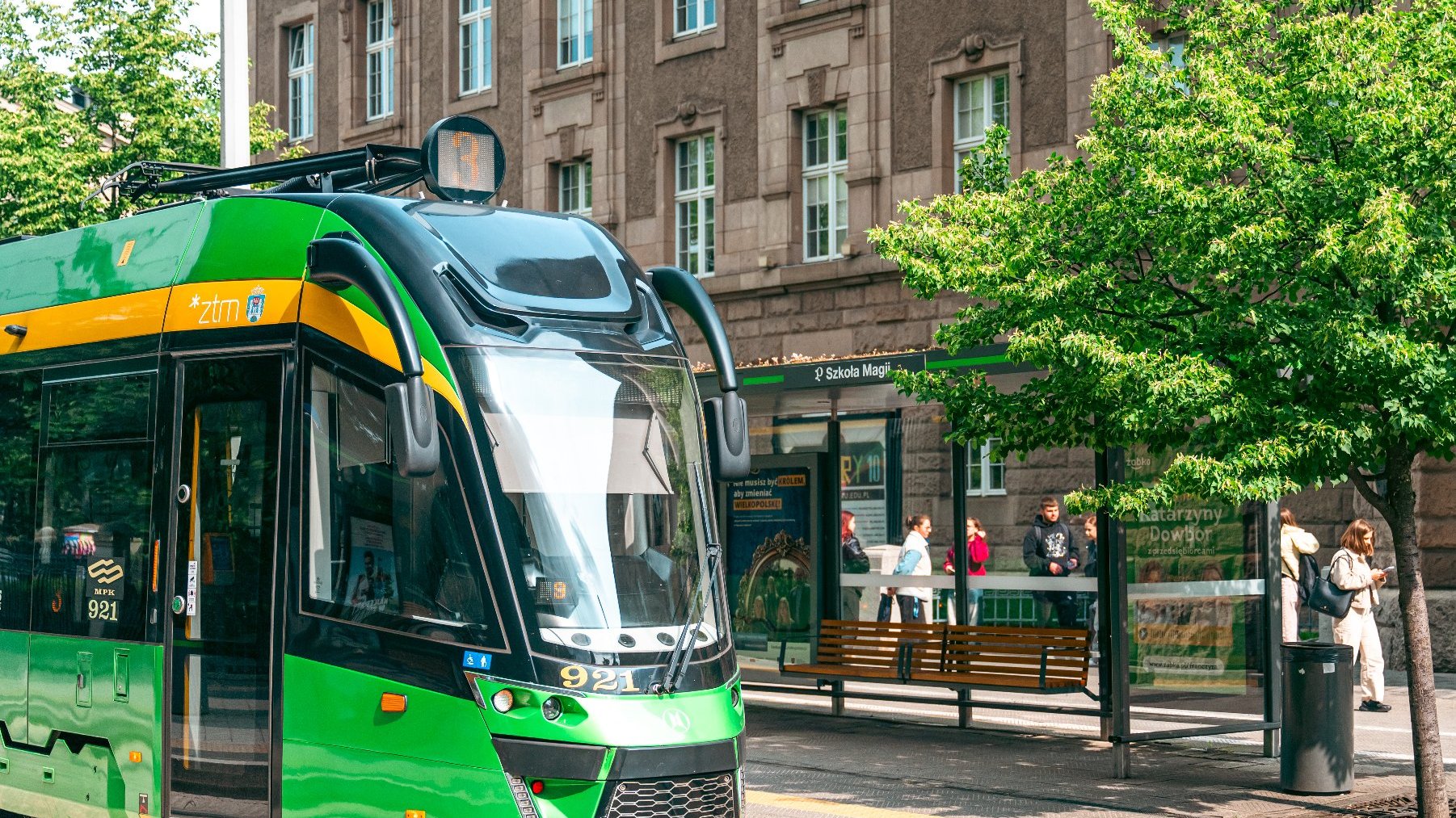 Zdjęcie przedstawia przystanek tramwajowy z napisem Szkoła Magii. Obok stoi tramwaj. Po chodniku chodzą ludzie.