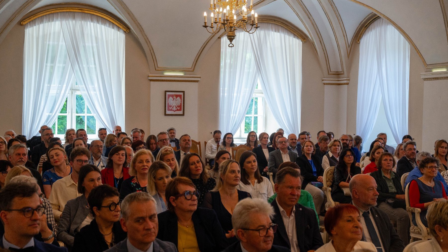 Duża grupa osób siedzi w eleganckiej sali, słuchając wykładu. Na ścianach widać wysokie okna z białymi zasłonami, a na suficie wisi ozdobny żyrandol. Na środku sali, między oknami, wisi obraz z godłem Polski.