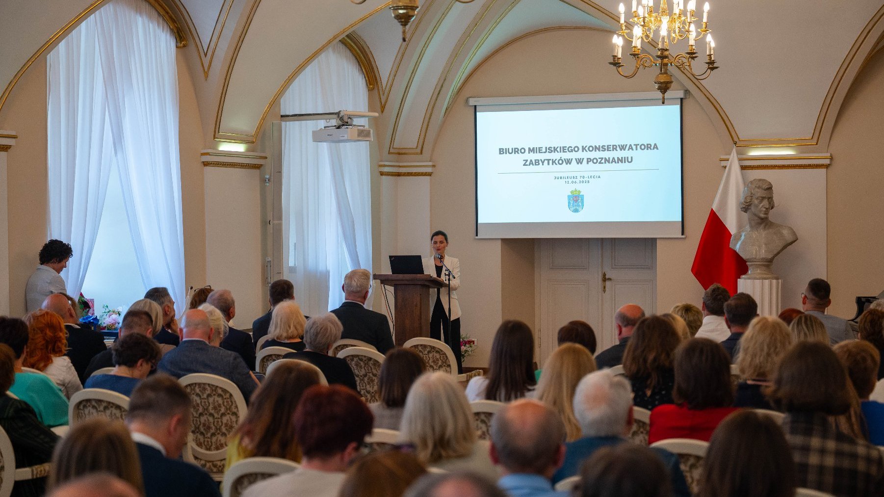 Kobieta w białej marynarce i czarnych spodniach przemawia do publiczności, stojąc za mównicą. W tle na ekranie wyświetla się prezentacja z napisem "BIURO MIEJSKIEGO KONSERWATORA ZABYTKÓW W POZNANIU" oraz logo miasta i data "JUBILEUSZ 70-LECIA 12.06.2025".