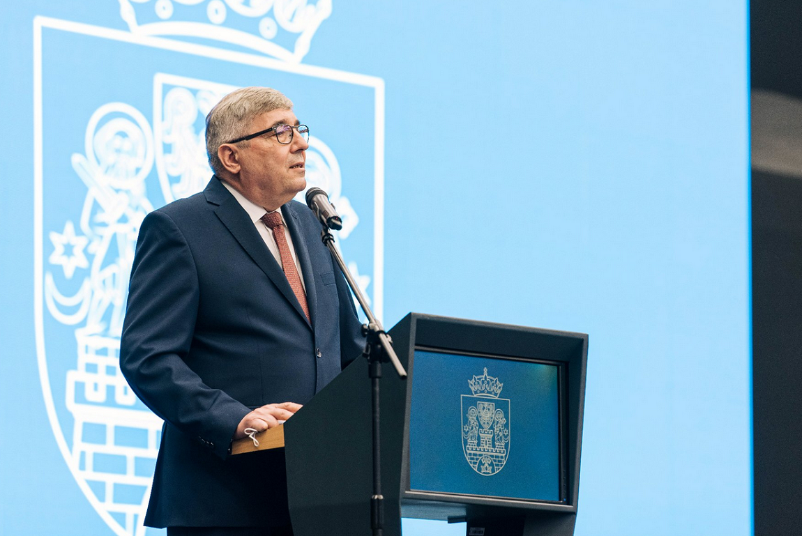 mężczyzna w garniturze, w okularach, o siewiejących włosach stoi przy mównicy z mikrofonem, na przodzie mówniu herb Poznania, za mężczyzą ściana z dużym herbem Poznania - grafika artykułu